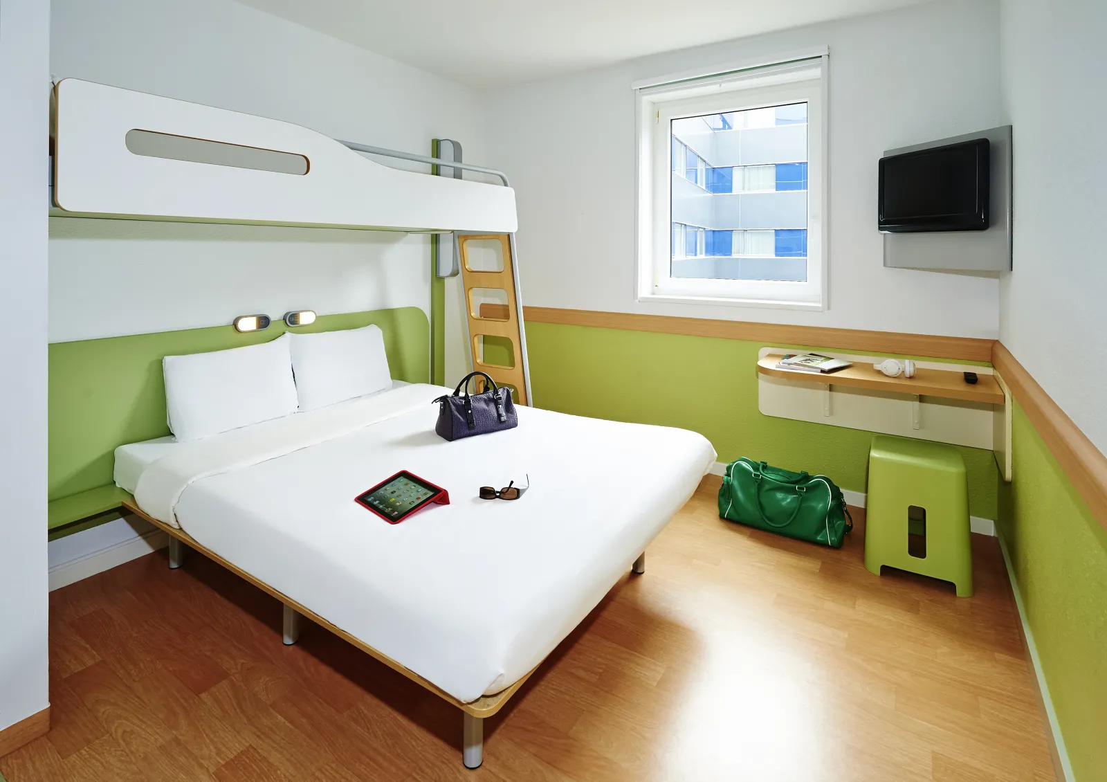 Modern hotel room at Ibis budget Bordeaux with white double bed, bunk bed, lime green and white walls, wall-mounted TV, window, and built-in storage.