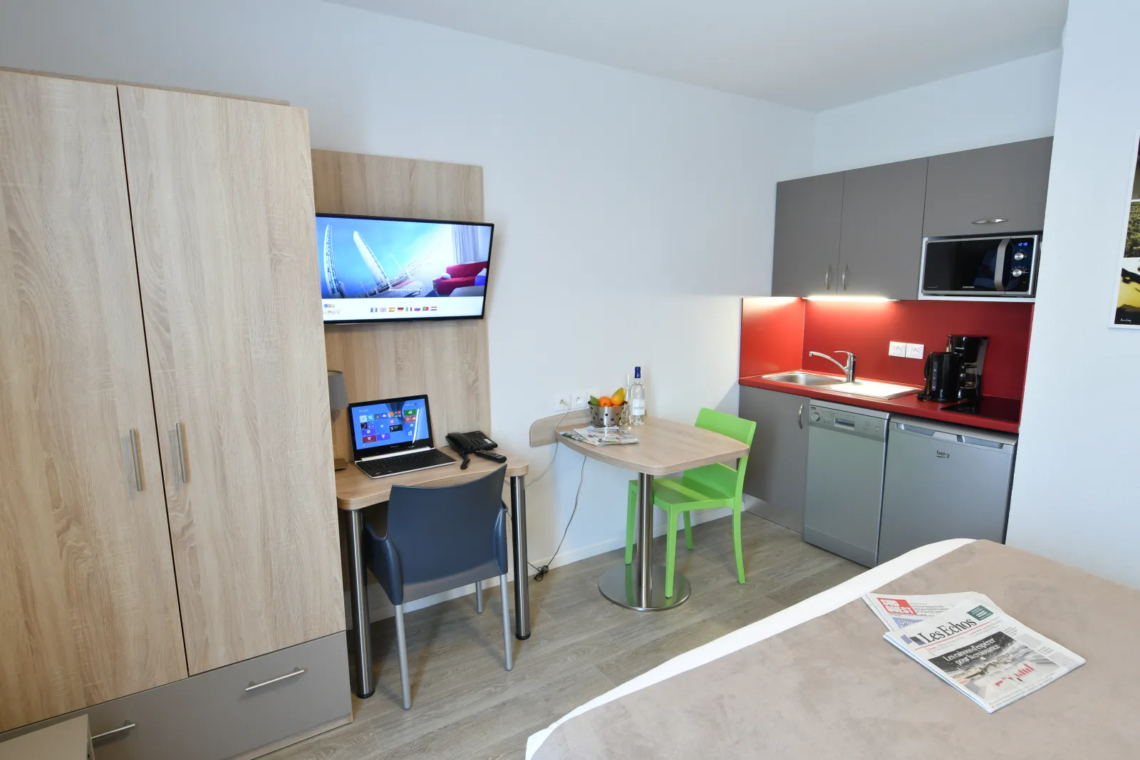 Modern studio apartment with equipped kitchen featuring red backsplash, desk area with laptop, dining table with green chairs, and light wood storage cabinets. Flat-screen TVs and microwave visible.