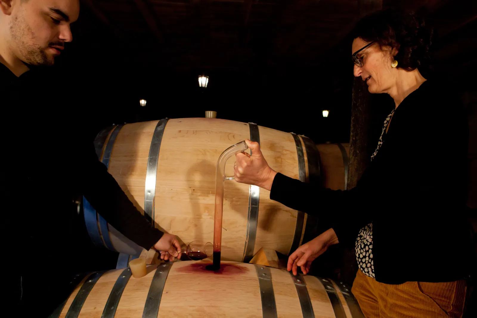 Two people inspect a wooden wine barrel bound with metal hoops in a dimly lit cellar with hanging lamps, in Bordeaux.