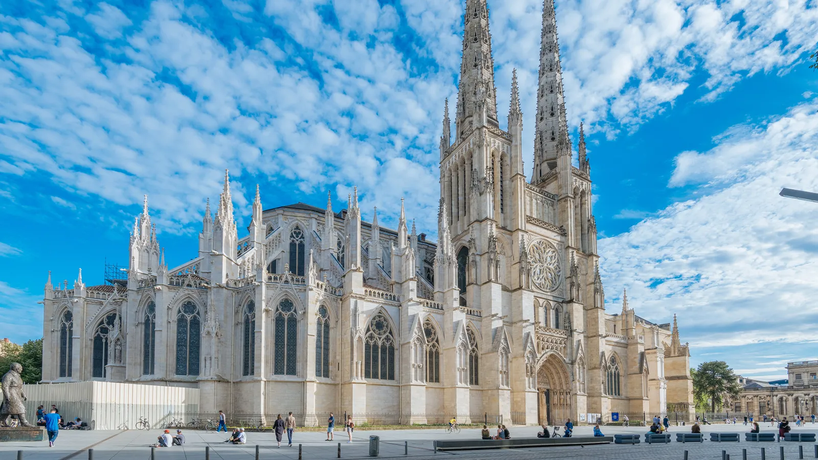 Bordeaux Cathedral of Saint-André, a white Gothic structure featuring two main towers, ornate facade with stained glass windows and rose windows, surrounded by visitors in a paved courtyard under a cloudy blue sky.
