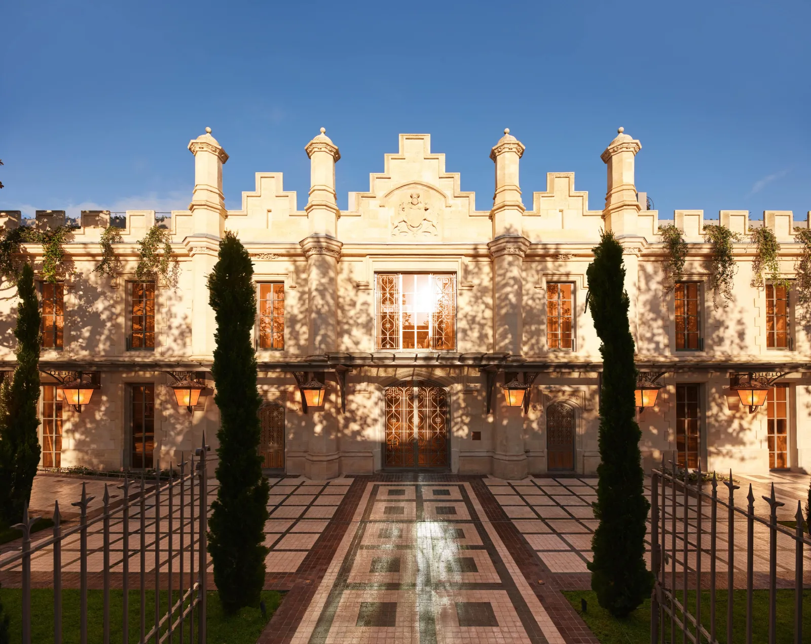 Facade of Mondrian Bordeaux Les Carmes, a historic Neo-Renaissance building featuring crenellations, turrets and ornate windows, framed by two green cypress trees and a paved courtyard in Bordeaux.