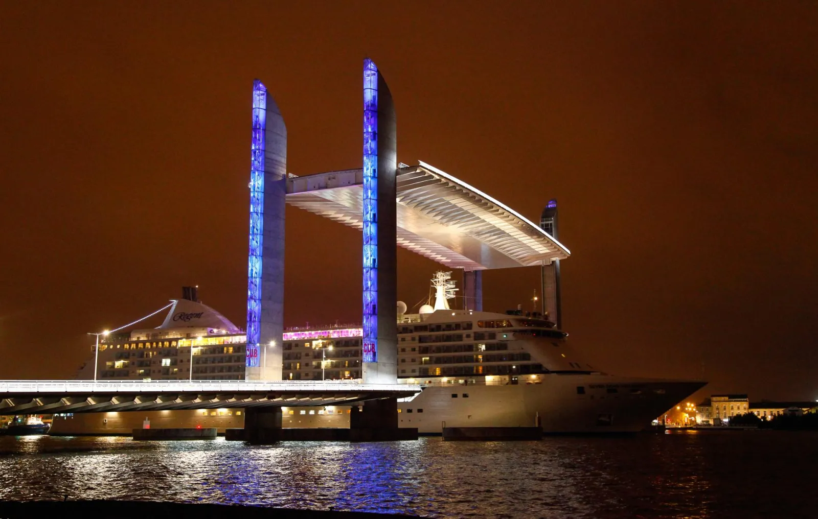 Bordeaux's Jacques Chaban Delmas Bridge illuminated at night, featuring its distinctive blue twin towers and raised deck spanning the Garonne River.