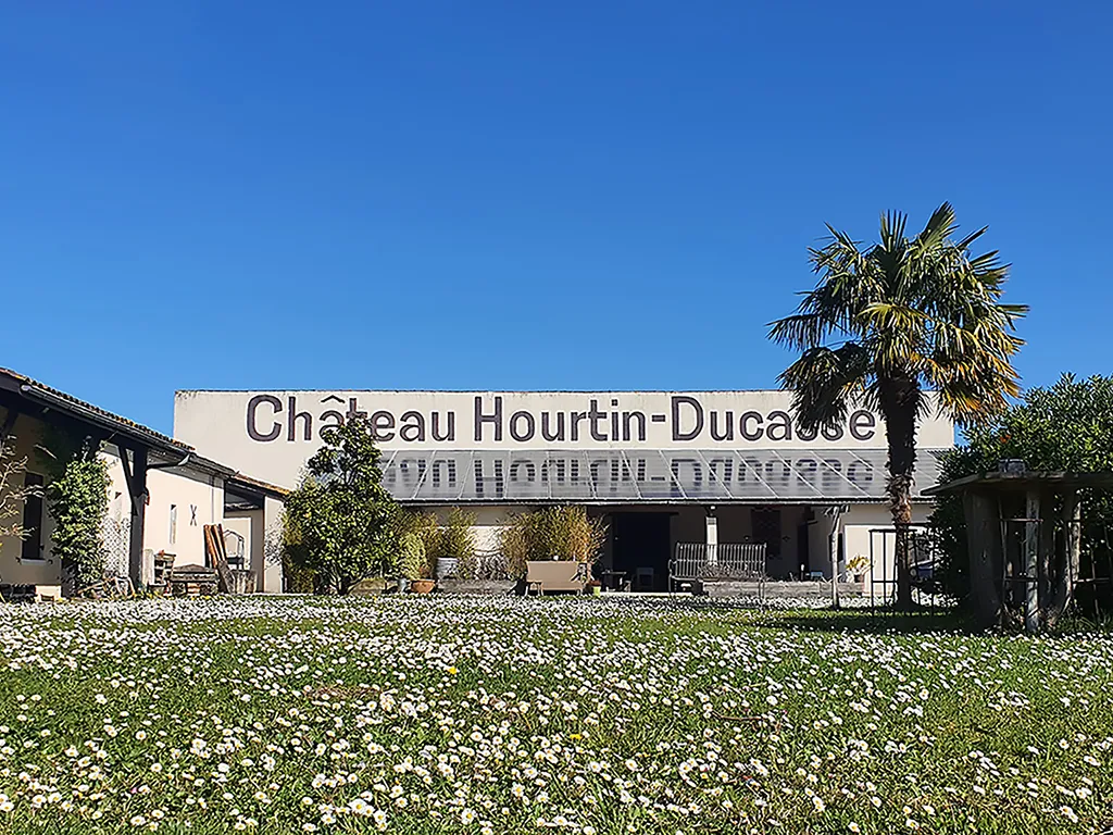Facade of Château Hourtin-Ducasse in Bordeaux with its name painted on the white wall, surrounded by a flowering garden and palm tree under clear blue sky.