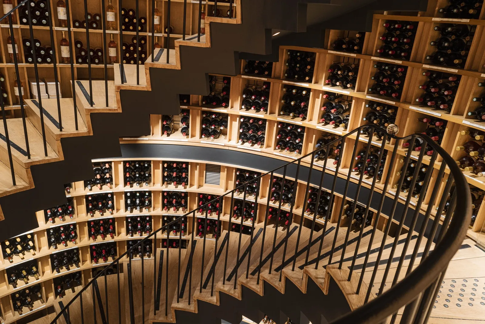 Spiral wine cellar with wooden shelves holding hundreds of wine bottles, black metal staircase winding through the space, in Bordeaux.