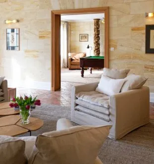 Elegant living room at Maison La Fleur de Boüard in Bordeaux featuring beige armchairs, exposed stone walls, red flower arrangement, and a view into a bedroom through large glass doors.