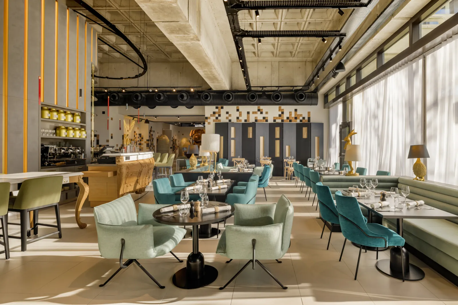 Contemporary restaurant interior featuring turquoise and pale green velvet chairs, black tables, gold lighting fixtures, bar area, and floor-to-ceiling windows with white curtains in Bordeaux.