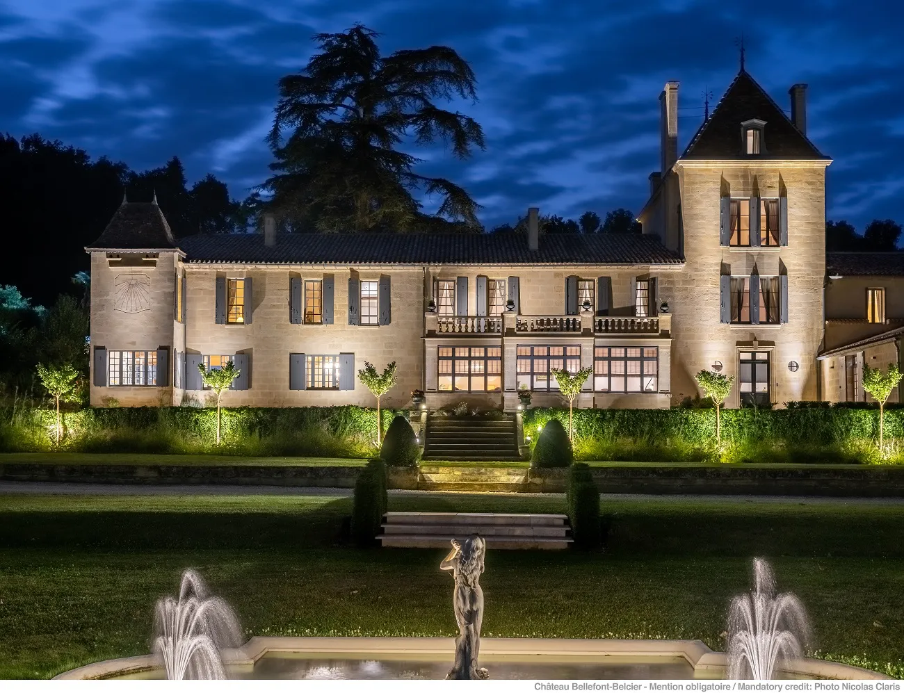 Château Bellefont Belcier in Bordeaux lit up at night, featuring a pale stone facade with pointed towers, central access stairs and illuminated fountains in the foreground