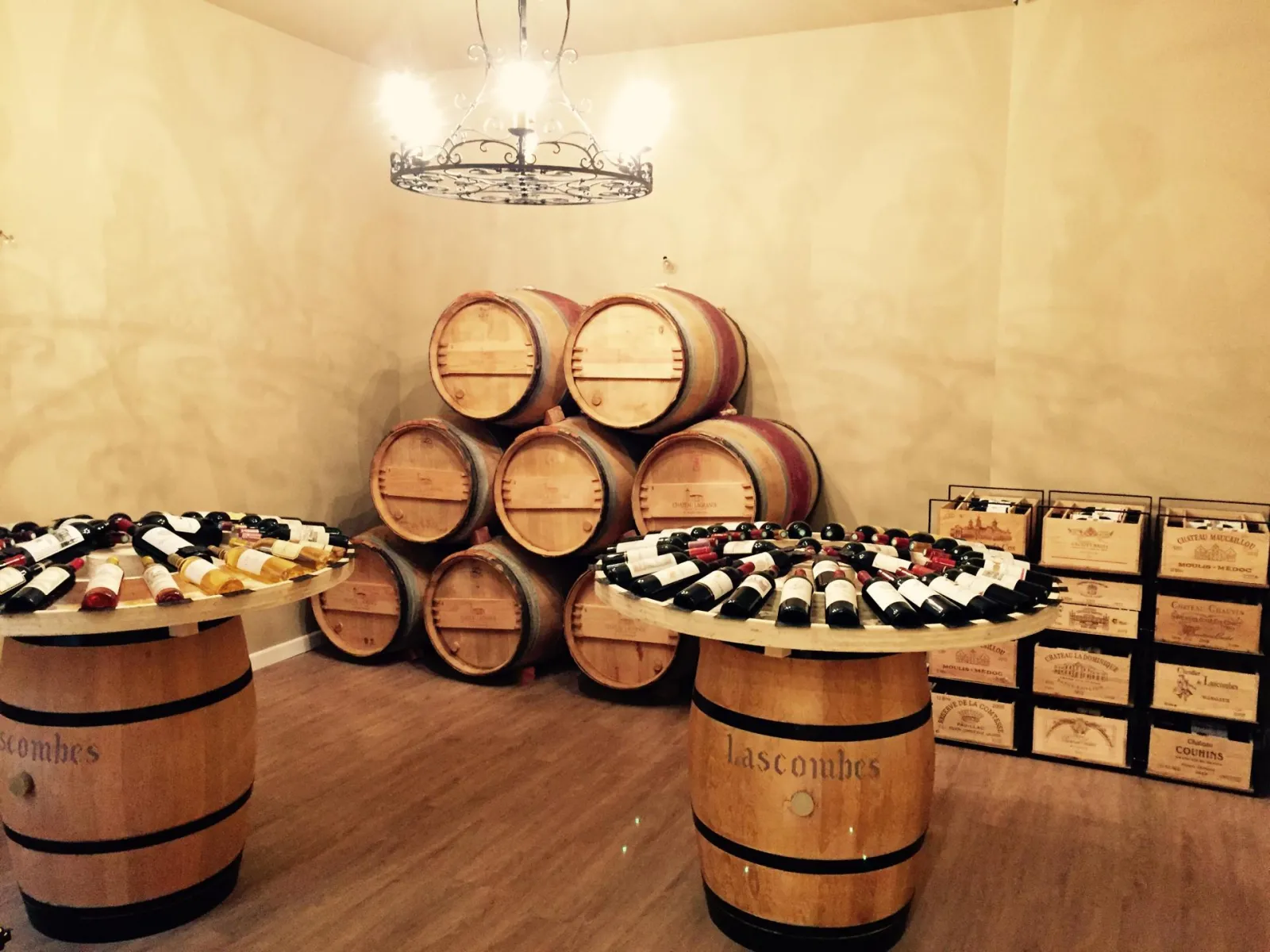Wine tasting cellar in Bordeaux featuring stacked oak barrels, wine bottles displayed on barrel tables, and storage racks, lit by an ornate iron chandelier.