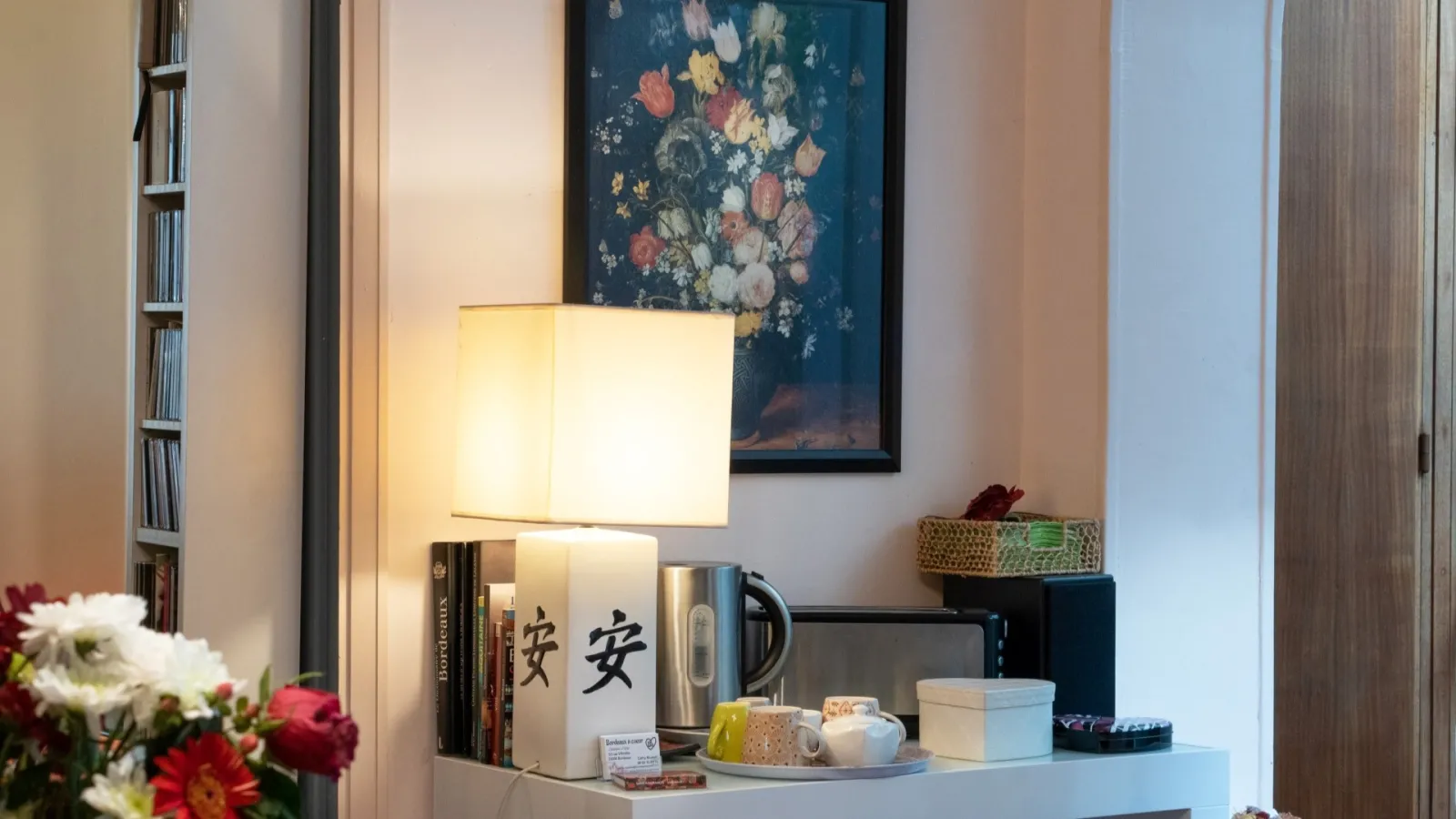 Elegant interior with table lamp, books, decorative mug with Asian characters, tray with accessories, fresh flowers in foreground, floral painting on wall and wall shelf with books