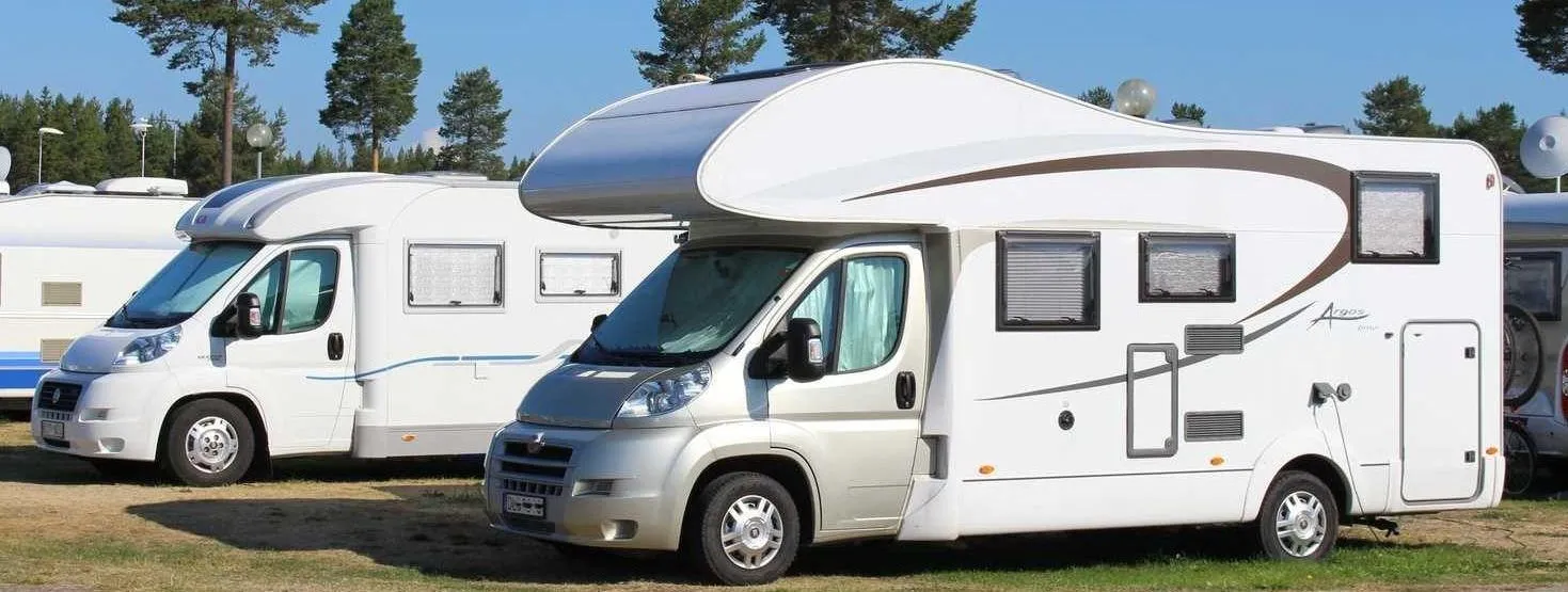 Three white motorhomes parked side by side in a parking area, with coniferous trees in the background and a clear blue sky.