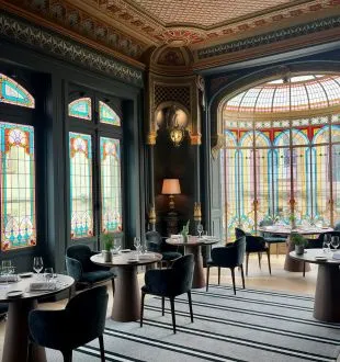 Elegant dining room at the Grand Barrail restaurant in Bordeaux featuring black furniture, ornate ceiling, colorful stained glass windows, and large windows overlooking the garden.