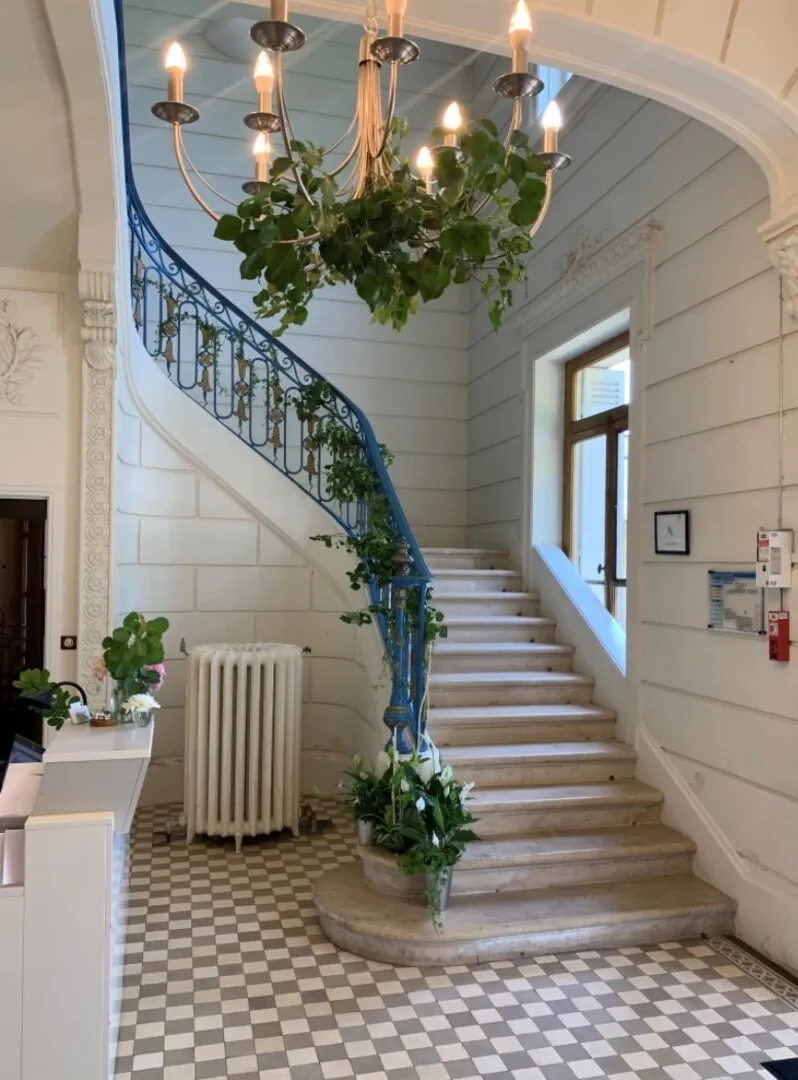 Elegant staircase at Château La Chèze in Bordeaux featuring a blue ornamental railing with ivy, suspended chandelier with green plants, black and white geometric tiling, and white walls.