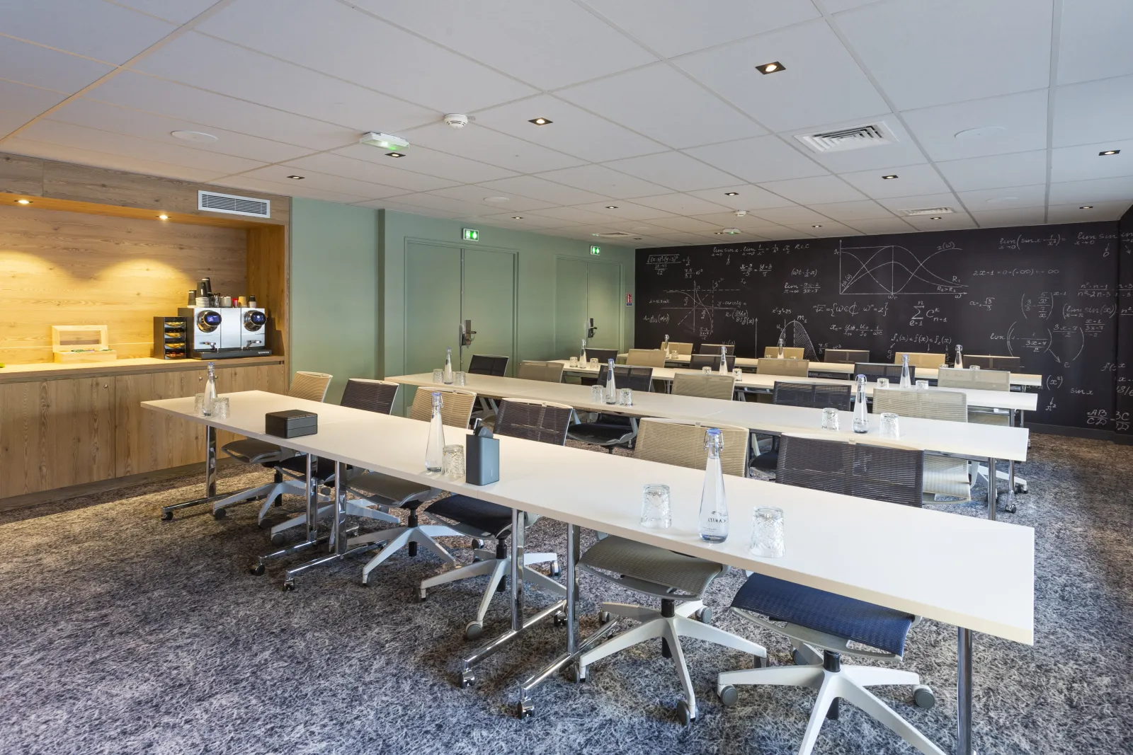 Modern meeting room at Novotel Bordeaux Centre featuring long white tables, black and beige chairs, blackboard with mathematical equations, yellow kitchenette, and mint green walls.