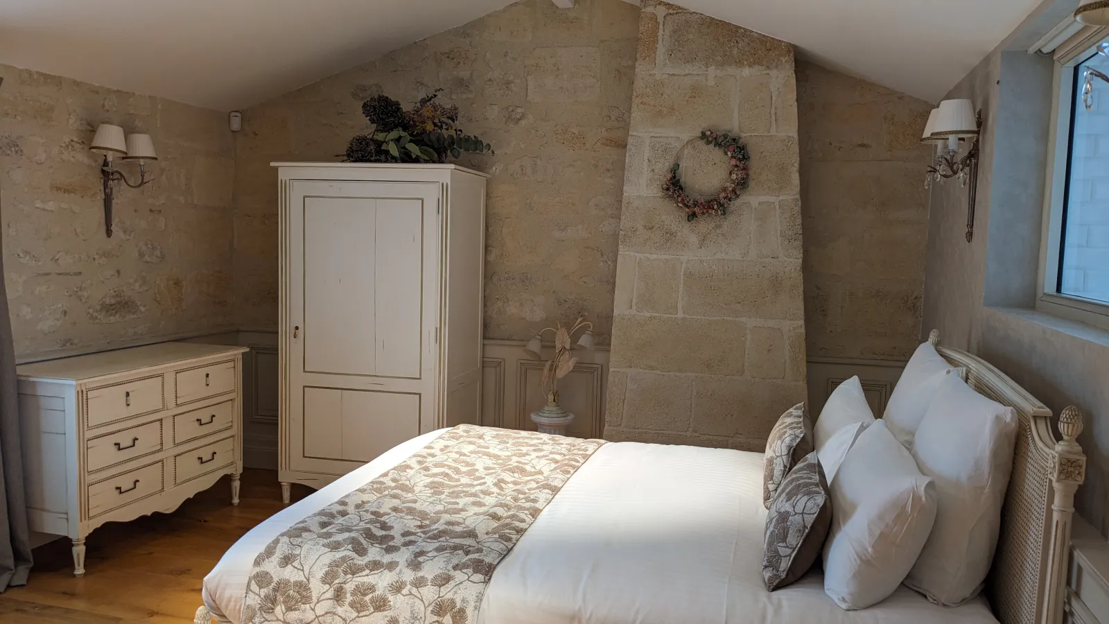 Elegant bedroom at Château La Rose Perrière in Bordeaux featuring exposed stone walls, king-size bed with white linens and patterned bedspread, cream-colored furniture, antique wardrobe, and rustic chic decor.
