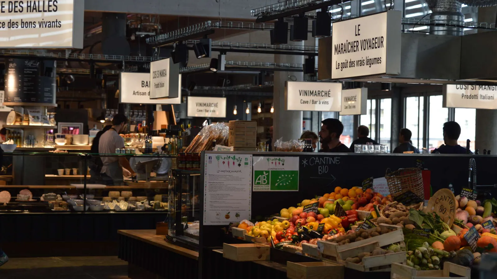 Interior view of Bacalan Market Hall in Bordeaux featuring organic fruit and vegetable stalls in the foreground, with shop counters and restaurant signage visible in the background.
