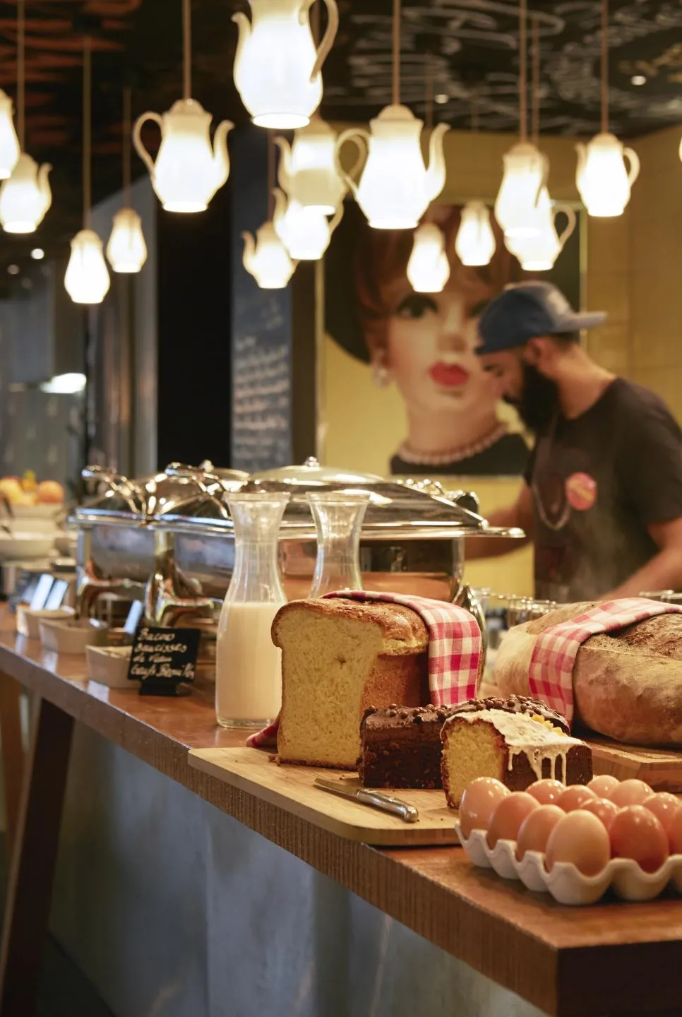 Bakery counter at Mama Shelter Bordeaux displaying breads, cakes and eggs, lit by decorative hanging lamps, with staff in the background
