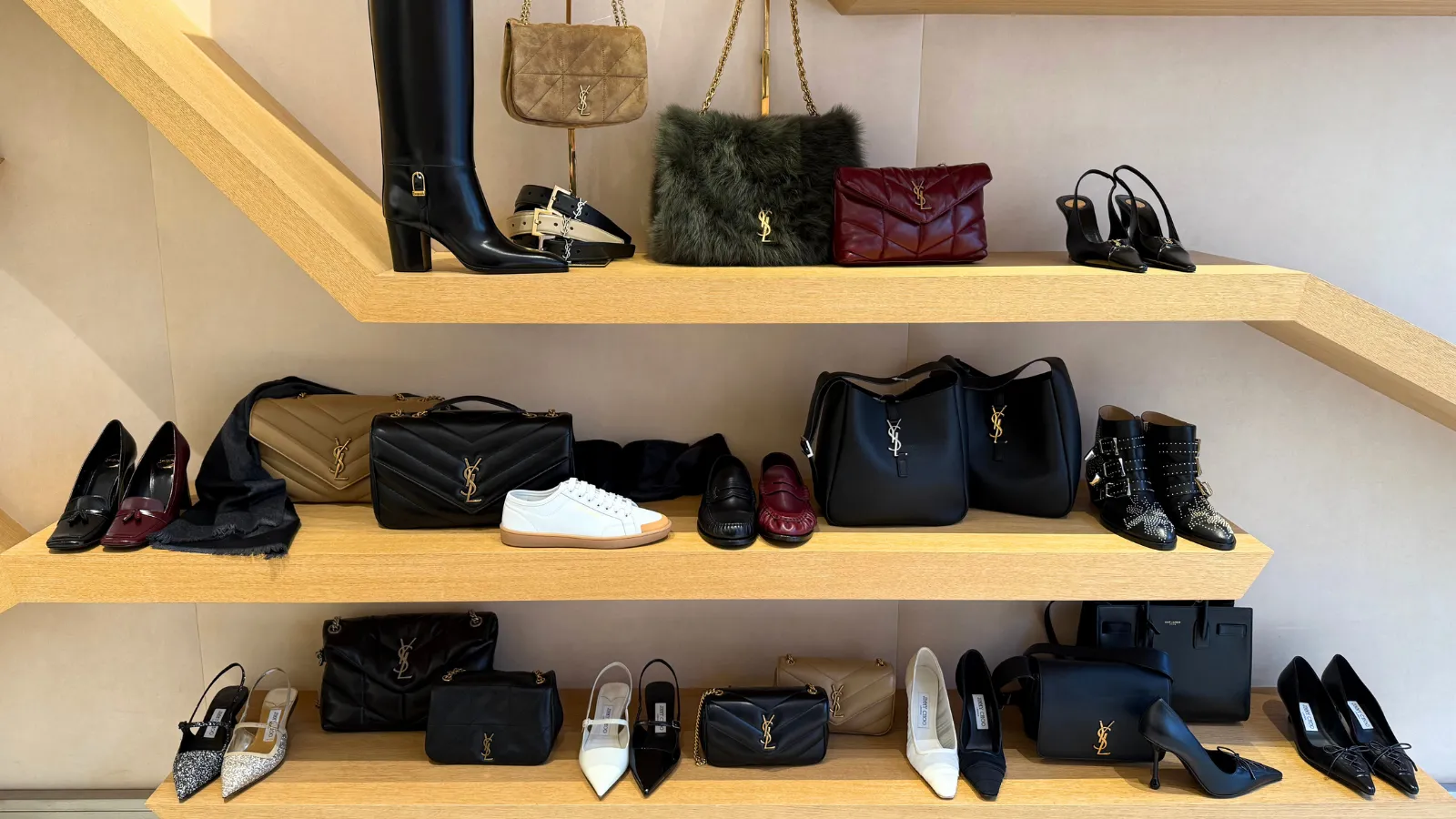 Three light wooden shelves displaying a luxury collection of shoes, handbags, and accessories including heels, sneakers, YSL bags, and ankle boots.