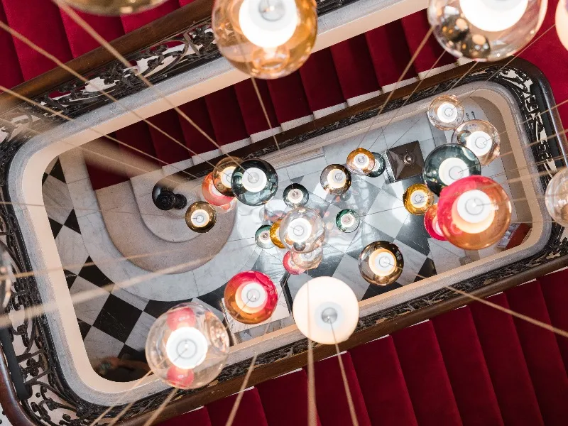 Overhead view of a luxury staircase at Hôtel Villas Foch in Bordeaux, decorated with colorful candles arranged on the steps and crystal chandeliers, featuring red walls and geometric black and white tiling.