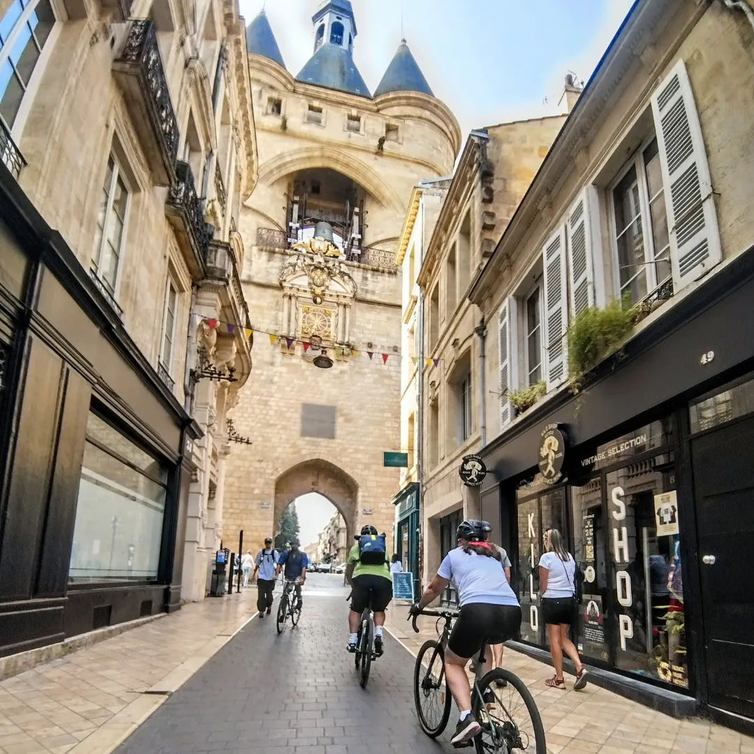 Paved street in Bordeaux with gravel bike riders passing in front of the Grosse Cloche, a historic medieval tower surrounded by stone buildings and shops.