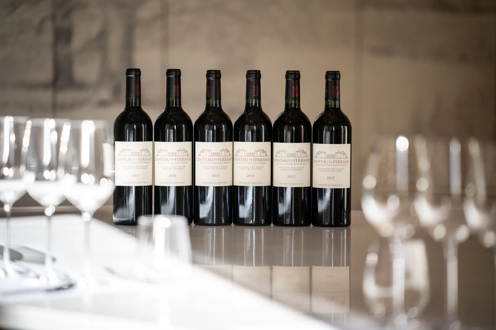 Six bottles of red wine from Château de Ferrand vintage years 2012 to 2017 lined up on a white table with blurred wine glasses in the background, in an elegant Bordeaux setting.