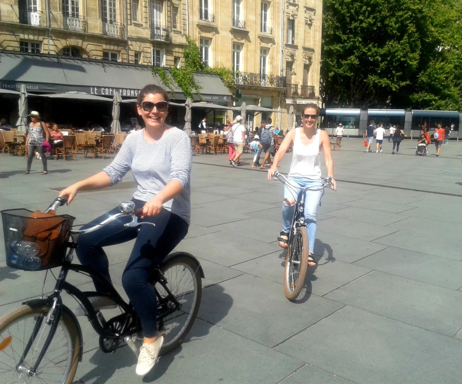 Two smiling women cycling on a paved square in Bordeaux, with Haussmann buildings and cafés in the background
