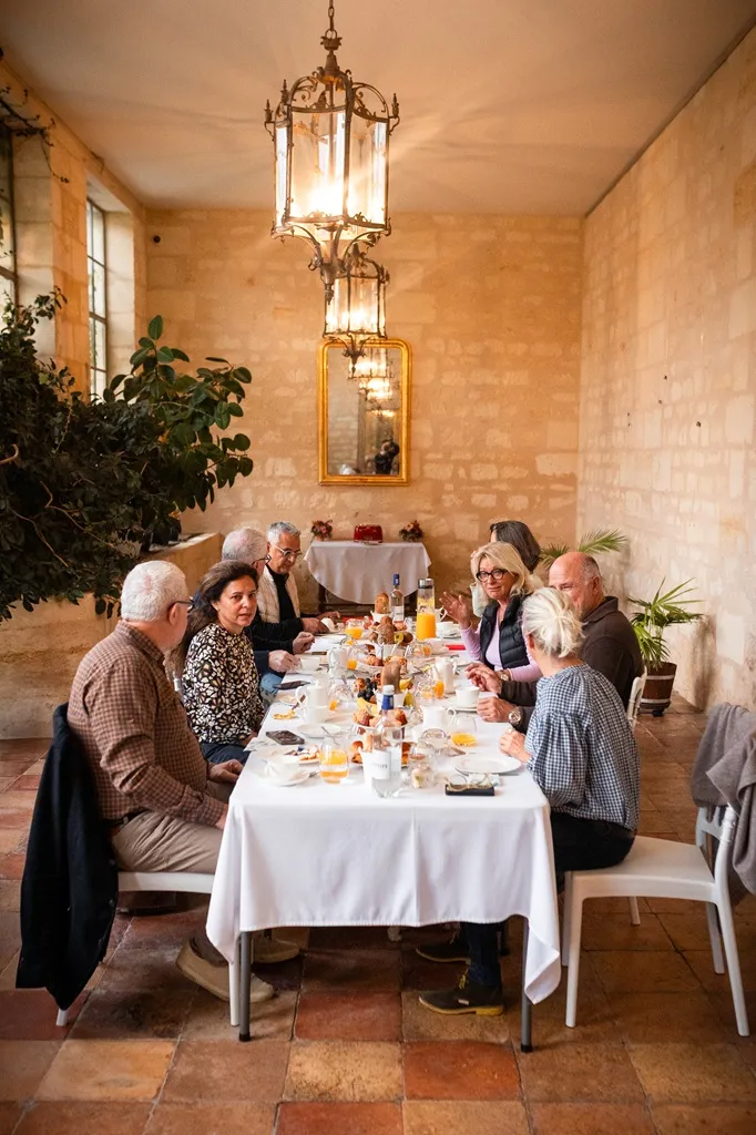 A group of elderly people sharing a meal around a round table in a stone room with ochre walls, lit by golden chandeliers.