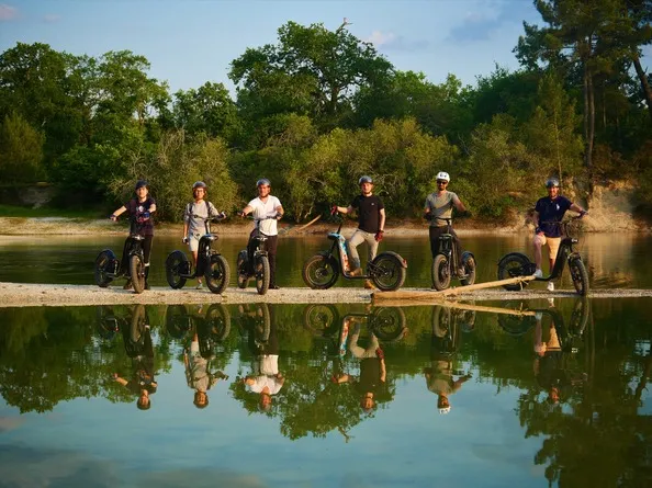 Seven people on bicycles and electric scooters lined up on a wooden bridge over a river, with green trees in the background and their reflections in the water.