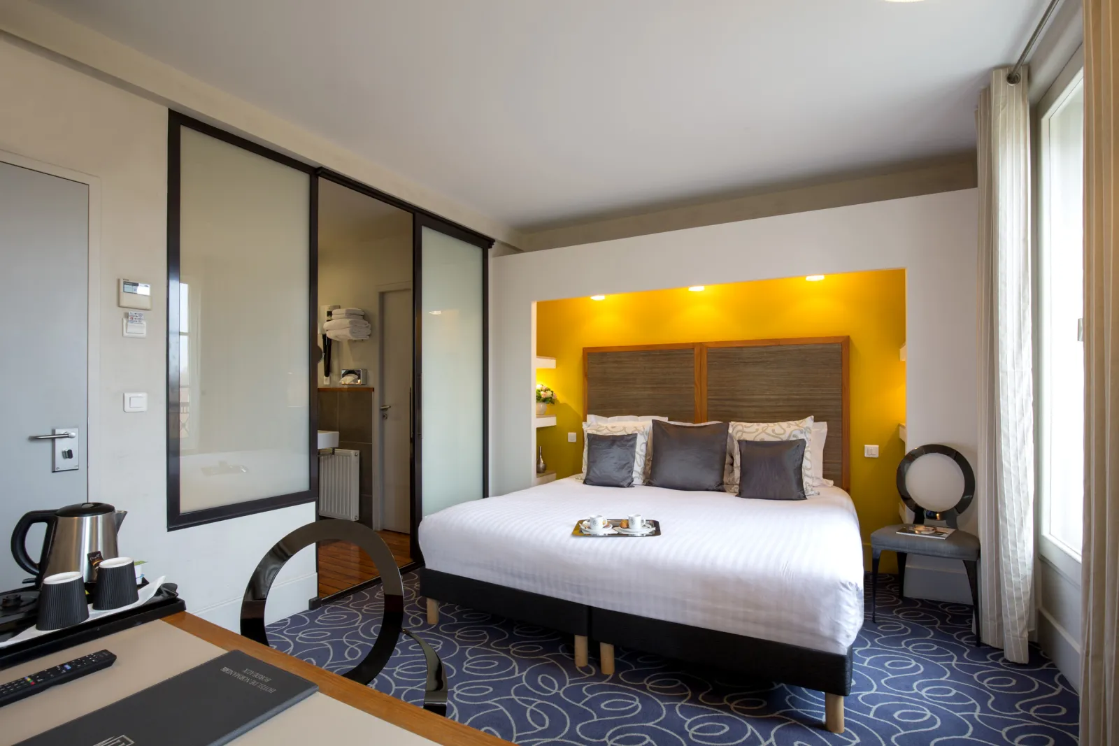 Modern hotel room featuring a queen-size bed with yellow accent wall, glass-enclosed bathroom, work desk with amenities, and contemporary décor in Bordeaux.