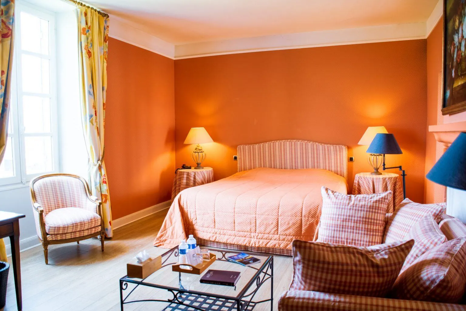 Elegant hotel room with vibrant orange walls, featuring a double bed with checkered headboard, two twin beds, bedside tables with lamps, a patterned armchair, and yellow floral curtains.