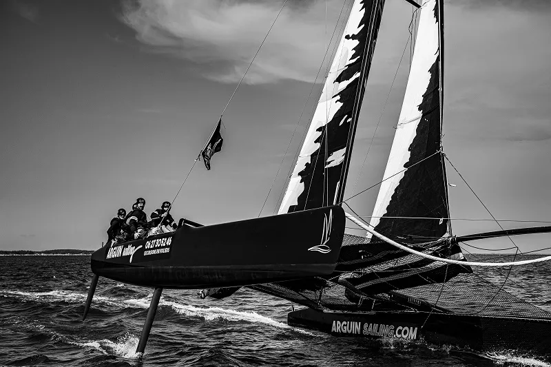 A black racing sailboat with crew sailing on water, featuring two white and black inflated sails, with Arguin Sailing name visible on the hull, photographed in black and white.