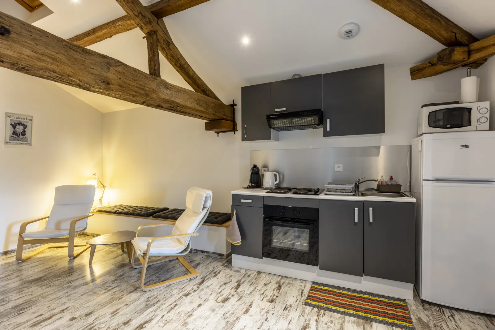 Studio Merlot at Château Pierre de Montignac in Bordeaux: equipped kitchen with grey units, relaxation area with beige armchairs, exposed wooden beams and polished concrete flooring.