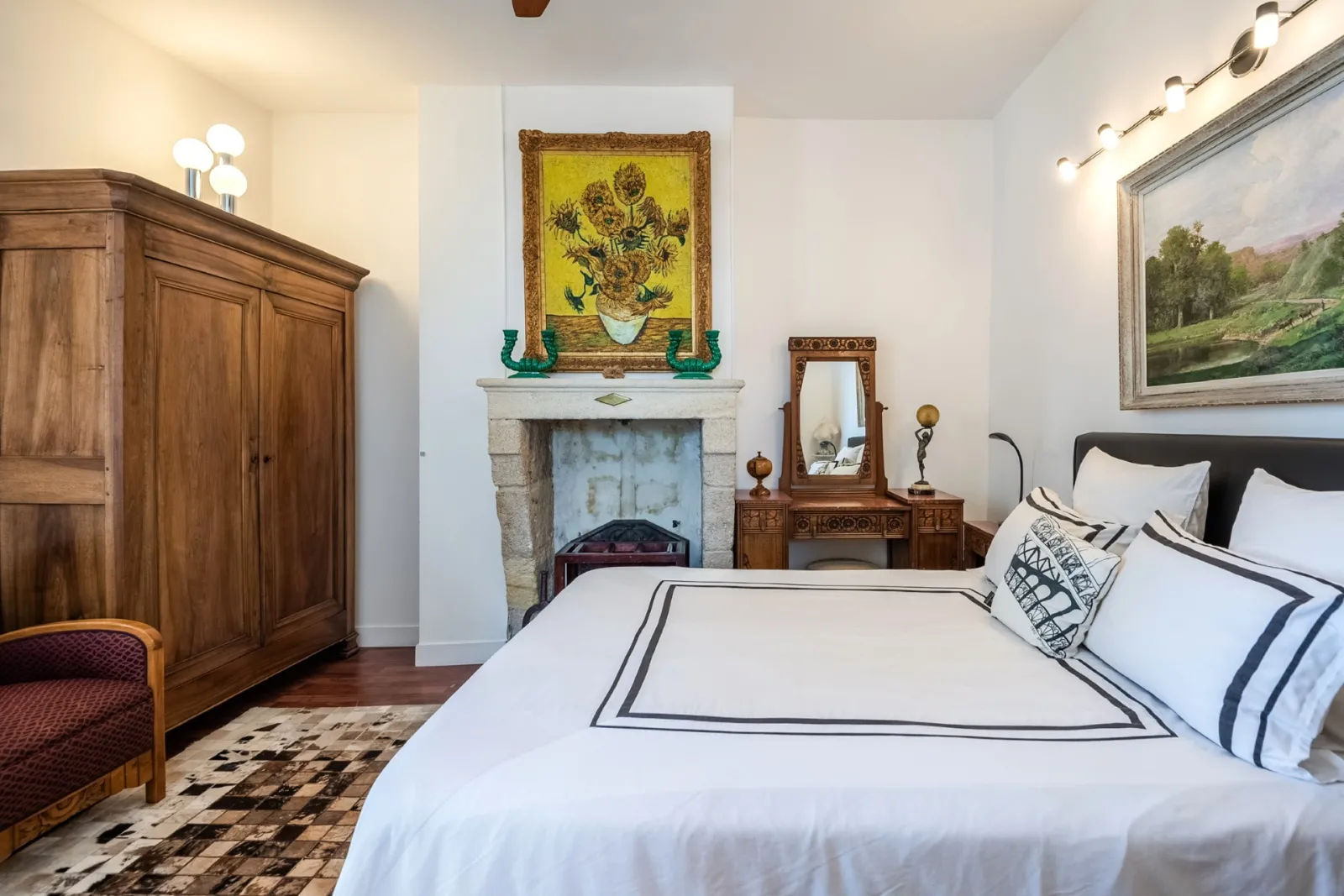 Elegant bedroom featuring a white bed with embroidered linens, solid wood wardrobe, decorated fireplace with sunflower painting, carved wooden dressing table, and landscape paintings.