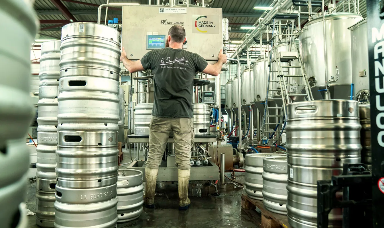 A brewer at Burdigala brewery in Bordeaux operates German Keg-Technik brewing equipment in a modern facility surrounded by metal beer kegs.
