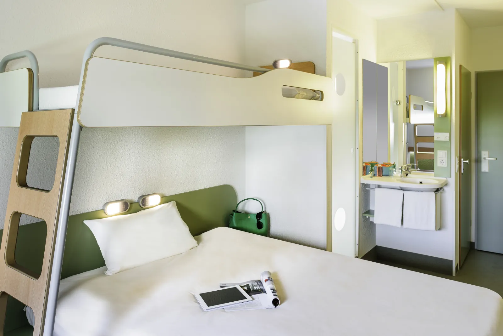 Budget hotel room featuring white and wood bunk bed with white bedding, green pillows, integrated reading lights, ensuite bathroom visible, and minimalist modern furnishings.