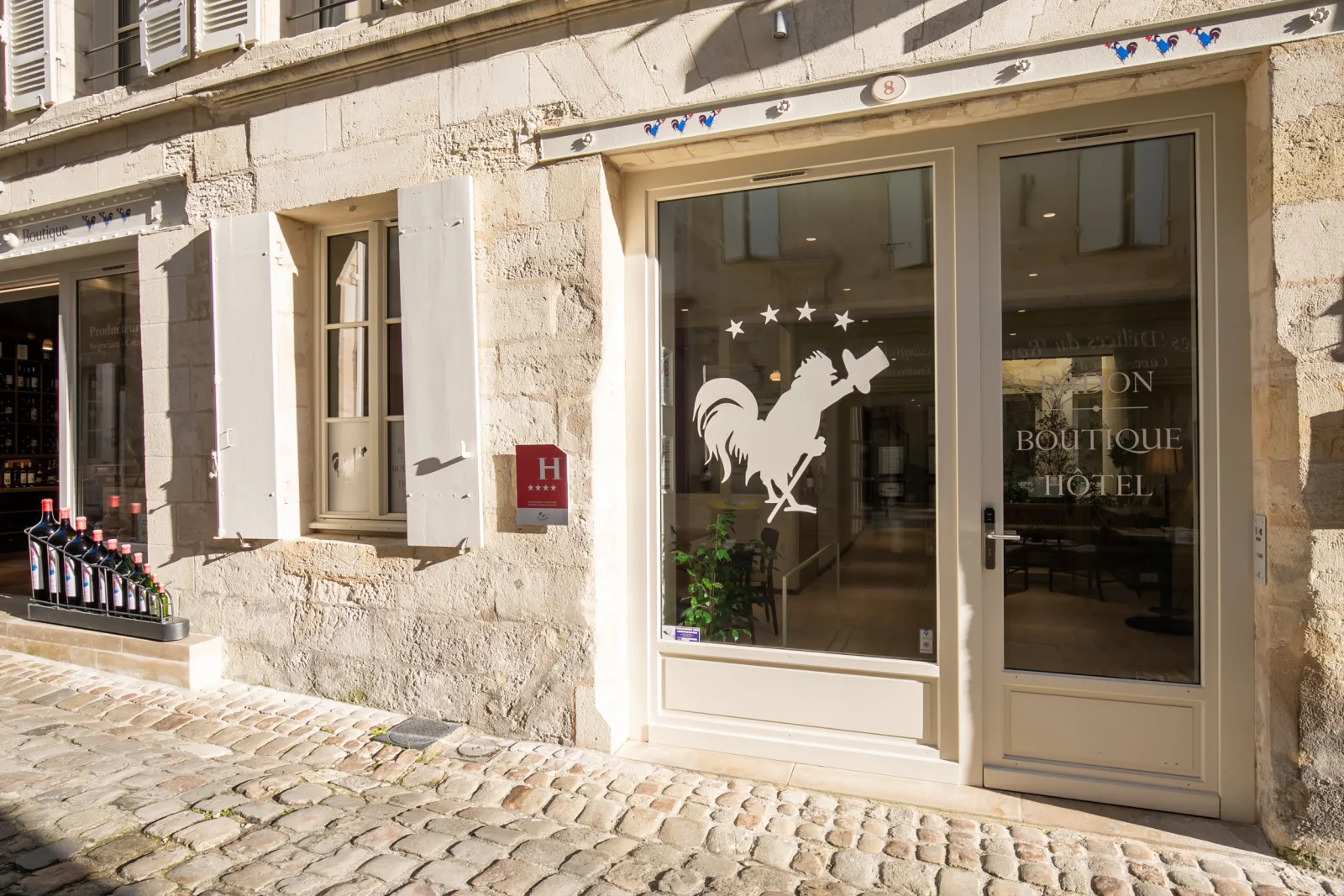 Storefront of Badon Boutique Hotel in Bordeaux featuring stone facade, white rooster logo, red hotel sign, and elegant display windows on a cobblestone street
