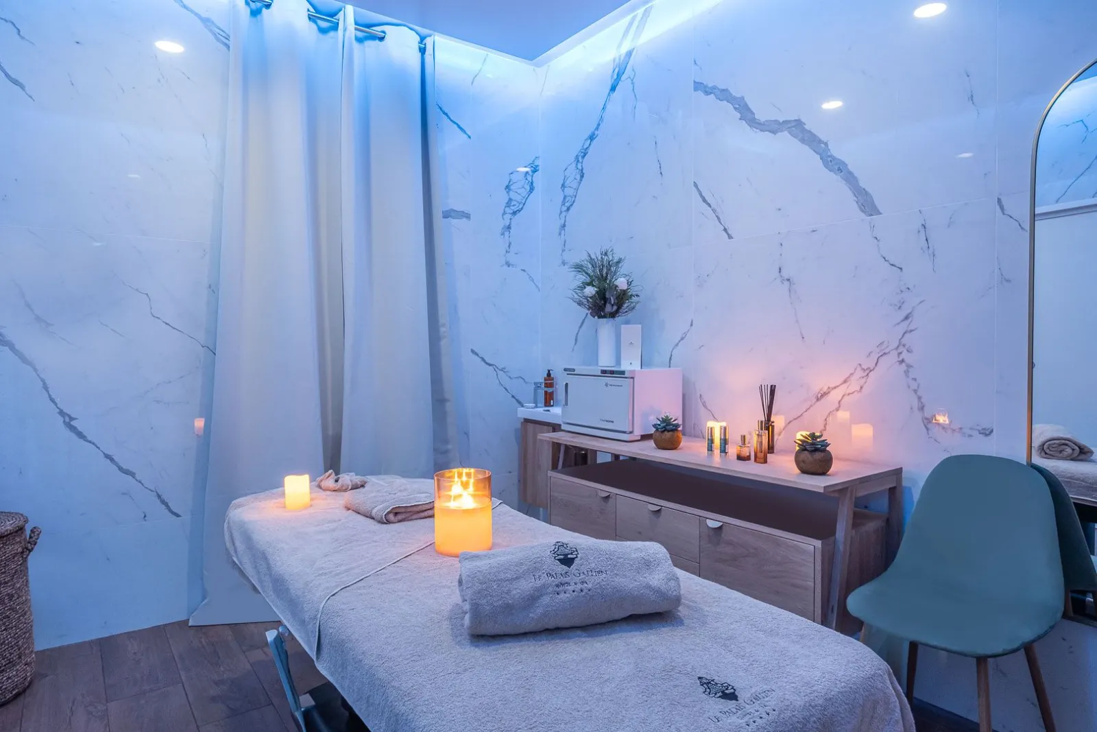 Luxurious spa treatment room with two cream-colored massage beds, lit candles, light blue marble walls, white curtains, green plants and wellness accessories at Palais Gallien Hotel & Spa in Bordeaux.