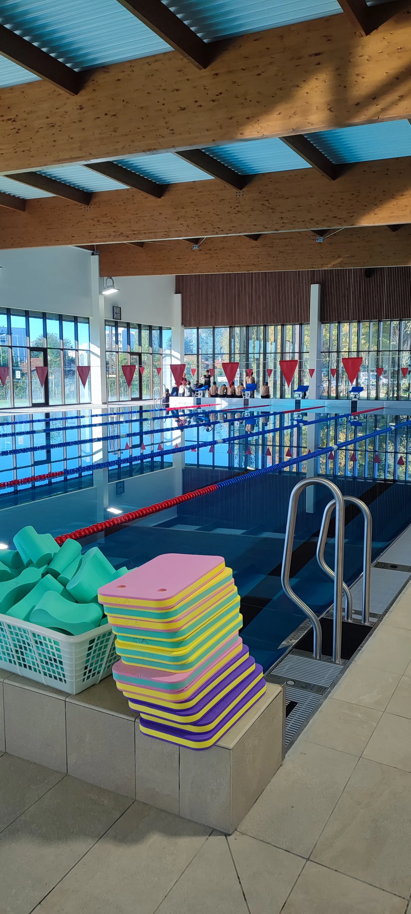 Indoor swimming pool with stacked colorful kickboards in foreground, blue water with lap lanes marked by red and white ropes, modern building with glass windows and perforated wooden ceiling at Elodie Lorandi complex in Bordeaux.