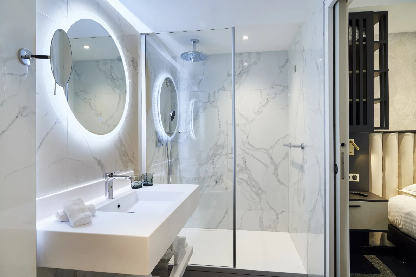 Modern bathroom featuring a white sink, illuminated oval mirror, walk-in shower with glass partition and white and gray marble wall cladding