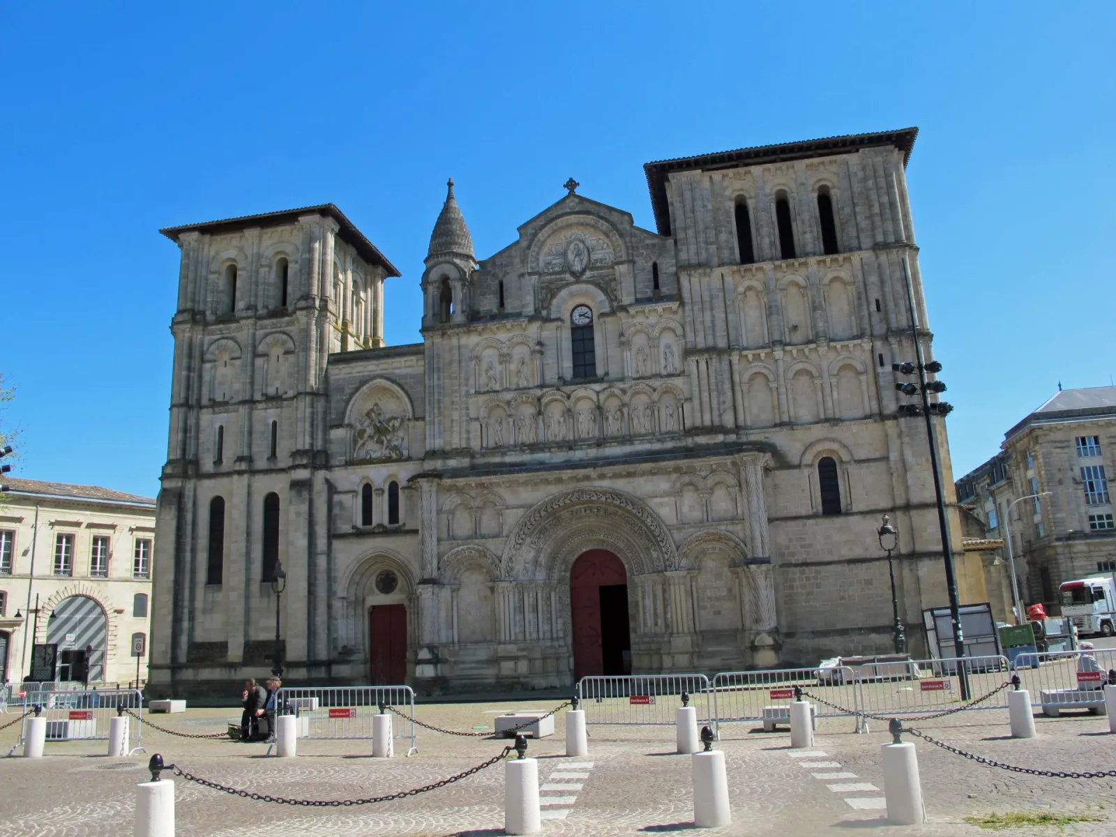 The Abbey Church of Holy Cross in Bordeaux, a Romanesque church with two square towers, ornate stone façade and rounded main portal, located on a paved square with safety barriers, under clear blue sky.