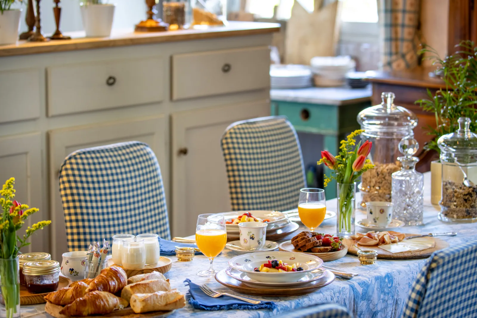 Elegant breakfast table set with croissants, orange juice, fresh fruits and flowers, blue checkered chairs, white furniture in the background in a guest room.