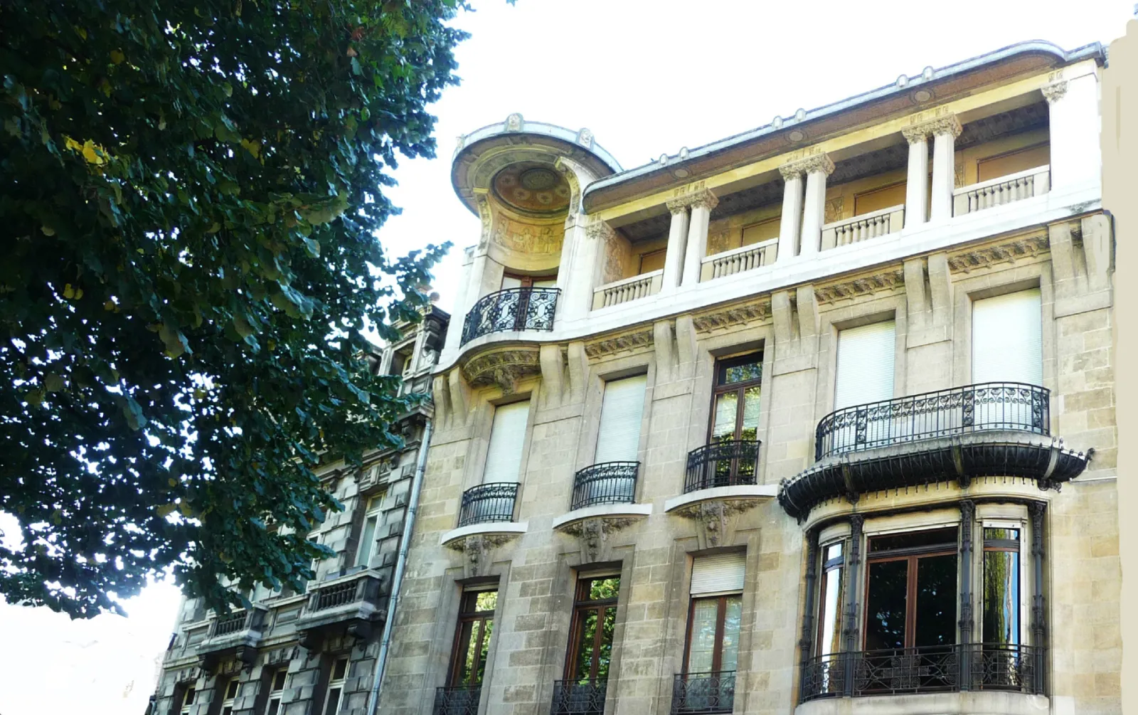 Elegant facade of the Frugès Hotel in Bordeaux, a Beaux-Arts building with stone masonry, wrought iron balconies and distinctive tower, surrounded by greenery