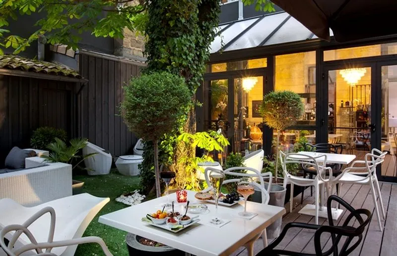 Elegant terrace of a boutique hotel in Bordeaux featuring white dining tables, modern chairs, landscaped garden, ambient lighting, and illuminated glass doors opening to the restaurant-bar interior.