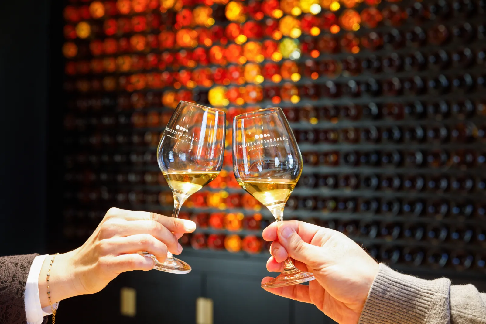 Two glasses of Sauternes raised in a toast against a backdrop of golden and orange bottles in a cellar at the Sauternes House in Bordeaux.
