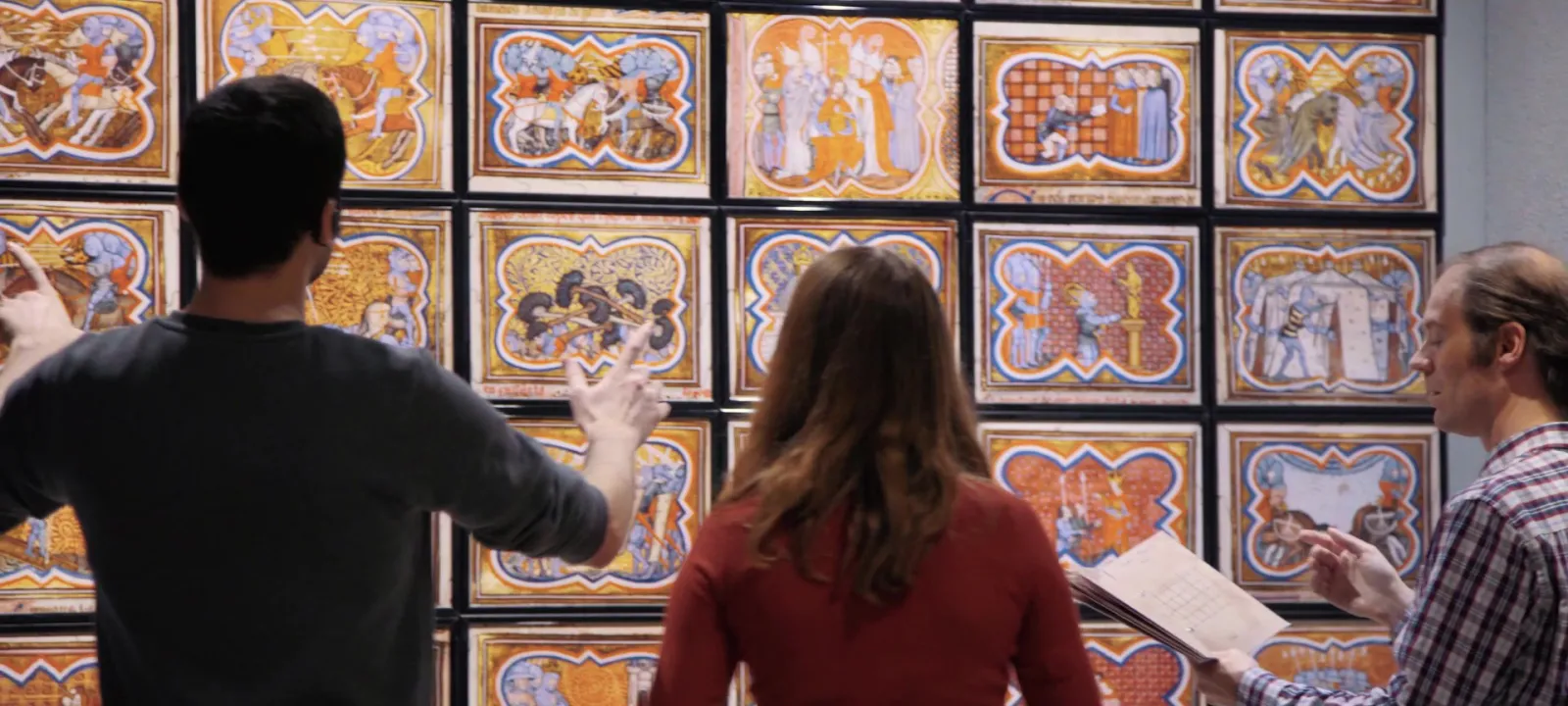 Three visitors examine a collection of ornate medieval ceramic tiles displayed at the Musée d'Aquitaine in Bordeaux, featuring hunting scenes and daily life with blue and orange geometric borders.