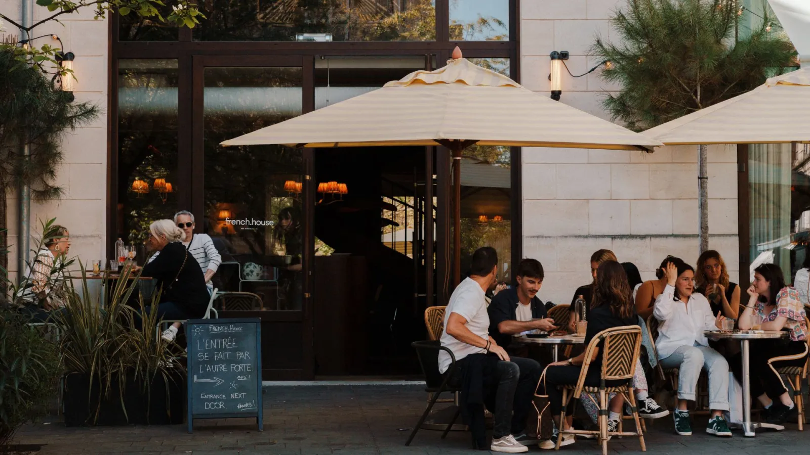 Outdoor terrace of French House restaurant in Bordeaux with customers seated under beige umbrellas, modern storefront with French signage