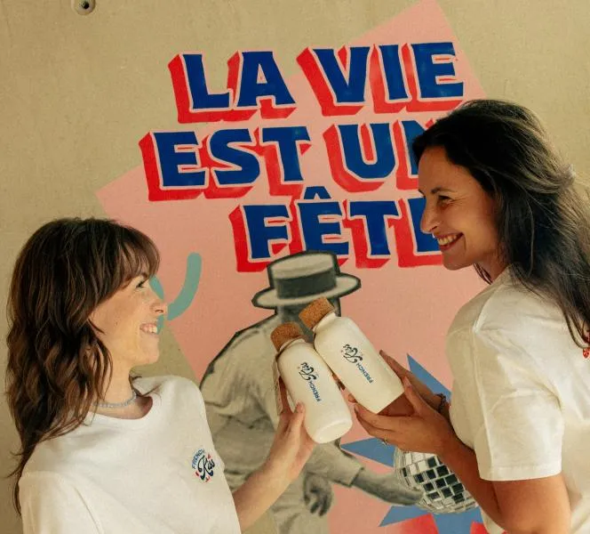 Two smiling women showcasing beauty products in front of a wall poster reading «Life is a celebration» in Bordeaux