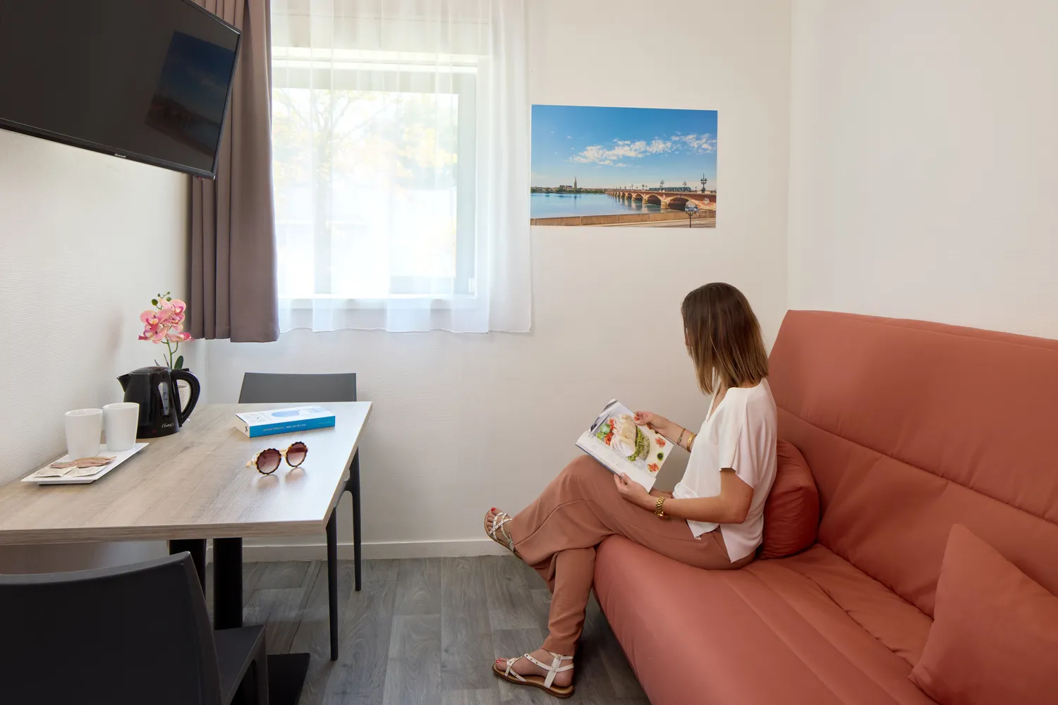 A woman sitting on a terracotta sofa in a modern room, holding a travel magazine, with a desk, bright window, and a photograph of Bordeaux on the wall.