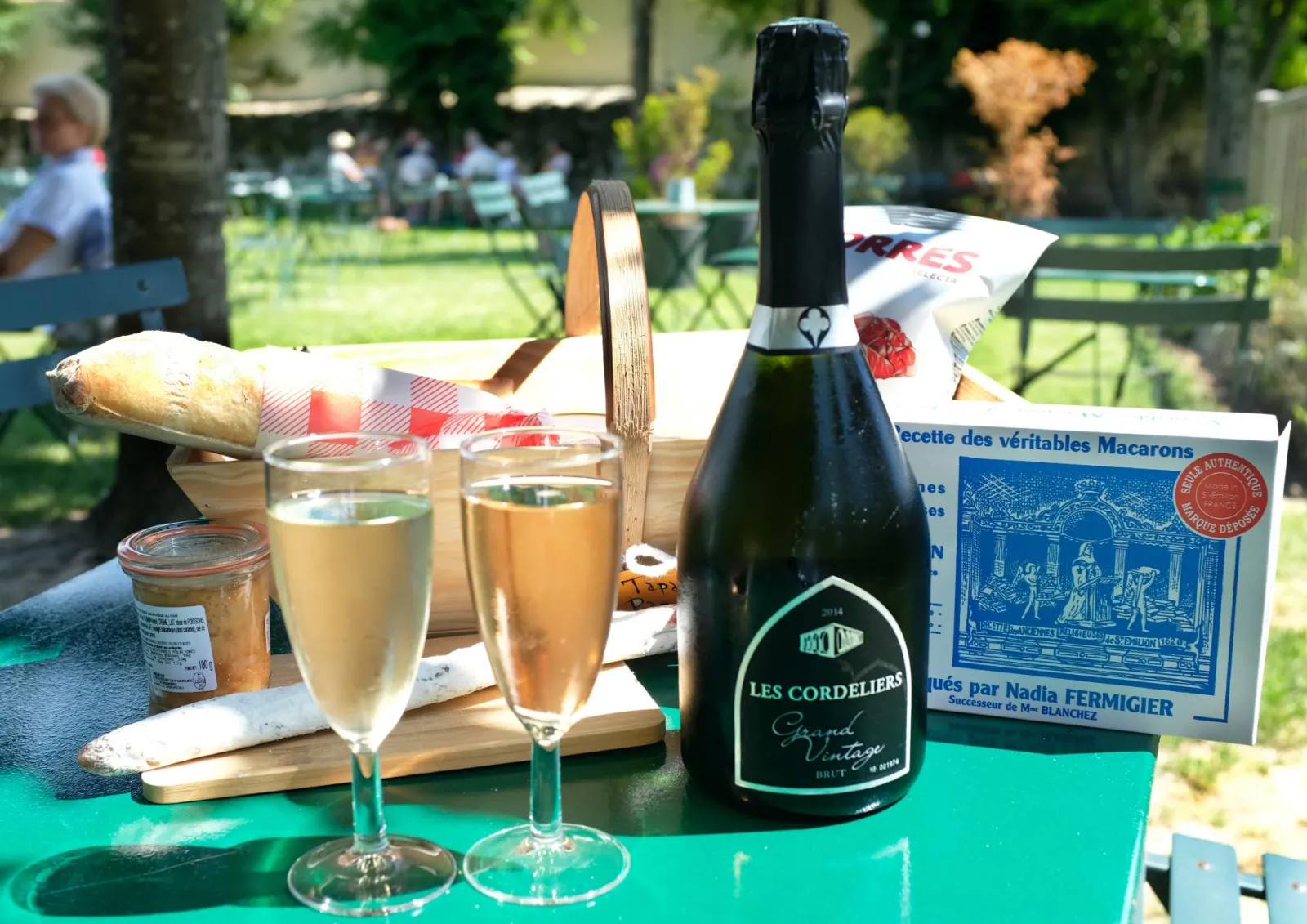 Picnic in a Bordeaux park featuring a bottle of Les Cordeliers champagne, two filled glasses, French bread, macarons and gourmet treats on a green table