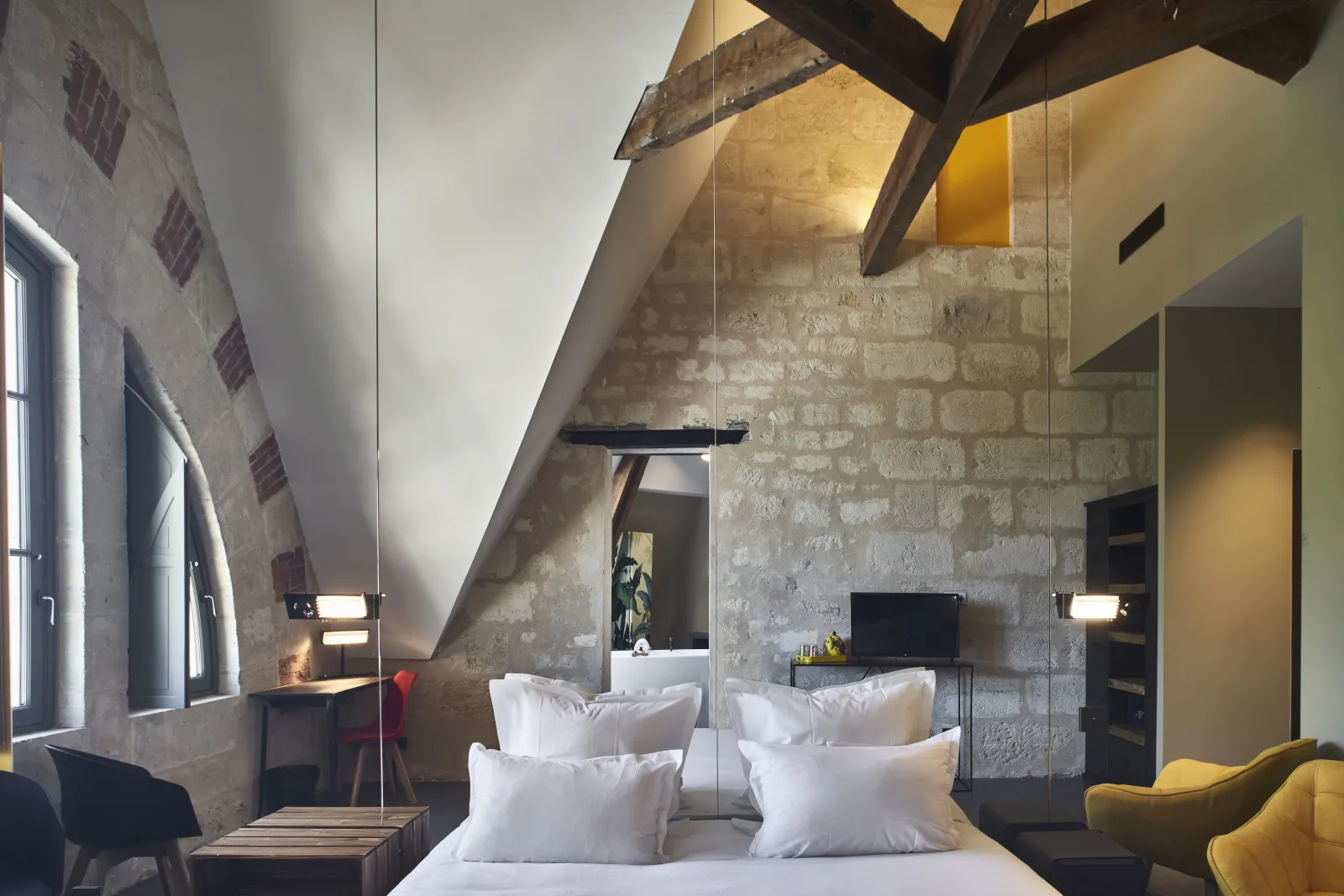 Modern attic bedroom at Hôtel La Zoologie in Bordeaux, featuring a white bed, exposed beams, raw stone walls, hanging lights, yellow accents, and contemporary furniture.