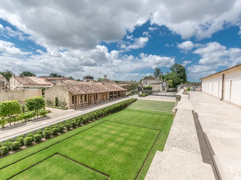 Aerial view of Château Marquis d'Alesme in Bordeaux featuring a manicured green lawn, stone buildings, and modern terrace under a cloudy blue sky